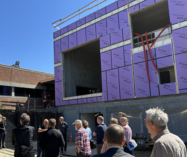 En gruppe mennesker står foran en rå bygningsfacade, mens en person taler.